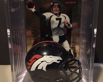 May include: A miniature Denver Broncos football helmet in a clear plastic display case with a trading card of John Elway. The helmet is black with an orange and white Bronco logo. The trading card is a gold foil card with a picture of John Elway in a Broncos uniform.