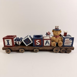 May include: A decorative wooden train with red, white, and blue blocks spelling "USA". A teddy bear wearing a hat and scarf sits on the train. The train has wheels and a star-shaped accent.
