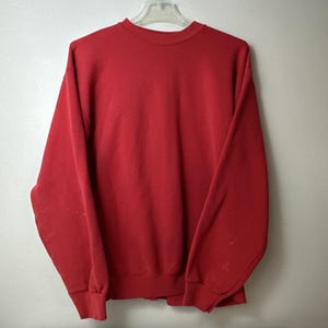 May include: A solid red long-sleeve crewneck sweatshirt featuring a ribbed collar, ribbed cuffs, and a ribbed hem. The garment is displayed on a clear plastic hanger against a plain, light-colored wall, showing a simple and classic design.