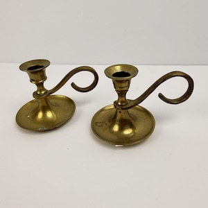 May include: Two vintage brass candle holders. Each holder has a circular base, a central cup for the candle, and a curved handle. The brass has a warm, aged patina. The candle holders are designed to hold a single candle.