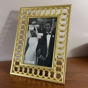 May include: A rectangular gold-toned picture frame featuring a decorative border of interlocking circular rings. The frame holds a black and white photograph of a wedding couple with text that reads Wilton Weddings and 4 in. x 6 in.
