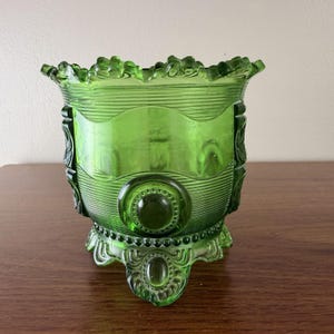 Könnte beinhalten: Eine grüne Glasschale mit dekorativem Design. Die Schale hat einen gerüschten Rand, horizontale Rippen und verzierte kreisförmige und ovale Akzente. Sie steht auf drei dekorativen Füßen.