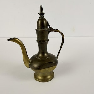 May include: A vintage brass pitcher with a curved spout and a handle. The pitcher has a bulbous body and a lid with a pointed top. The brass has a two-tone finish, with a lighter color on the lower portion and a darker color on the upper portion.