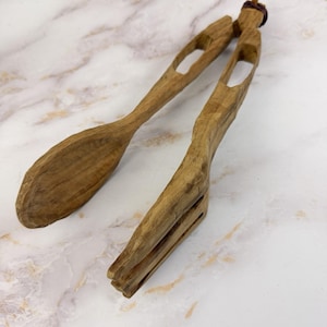 Puede incluir: Juego de cuchara y tenedor para servir ensalada de madera. La cuchara tiene una cabeza ancha y ovalada, y el tenedor tiene tres púas. Ambos utensilios tienen una veta de madera natural y un agujero en el mango. El juego es de madera marrón claro.