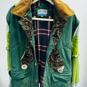 May include: A green jacket with a mustard yellow corduroy collar, featuring dark paisley lapels, a plaid interior lining, and sleeves with green tree graphics and textured knit sections. The label reads Northeast Terrain Company.