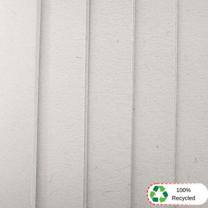 May include: A stack of light gray, textured cardboard sheets arranged vertically. The surface of the material appears fibrous and matte. In the bottom right corner, a white rectangular label features a green recycling symbol and the text 100% Recycled.