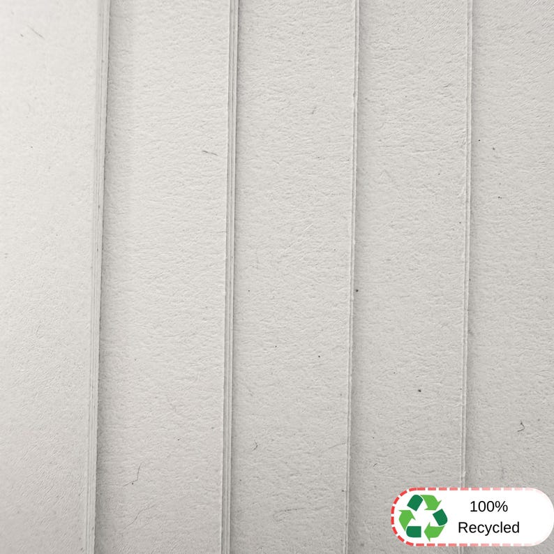 May include: A stack of light gray, textured cardboard sheets arranged vertically. The surface of the material appears fibrous and matte. In the bottom right corner, a white rectangular label features a green recycling symbol and the text 100% Recycled.