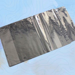 May include: A rectangular, metallic grey wallet with a reflective surface. The wallet appears to be made of a flexible material and has a slightly textured appearance. The wallet is set against a light blue background with a topographic map design.