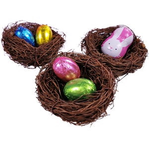 May include: Three brown twig nests containing colourful foil-wrapped chocolate eggs and a pink chocolate bunny with a white face.