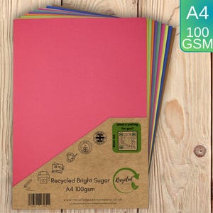 May include: A stack of A4 100 GSM recycled bright sugar paper in various colours, including pink, orange, yellow, green, blue, and brown. The pack features a brown paper wrapper with icons, a QR code, and the text Recycled Bright Sugar A4 100gsm.
