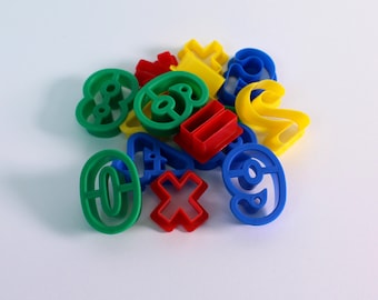 Könnte beinhalten: Eine Sammlung bunter Plastik-Ausstecher in Form von Zahlen, Buchstaben und Symbolen. Die Ausstecher sind in den Farben Grün, Blau, Rot und Gelb.
