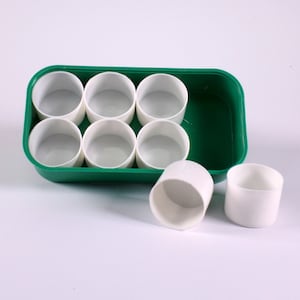 Op de afbeelding: Een groene rechthoekige bak met zes witte, cilindrische bekers. Twee extra bekers staan buiten de bak. De bekers hebben een gladde, glanzende afwerking en zijn leeg. De bak en bekers staan op een witte ondergrond.