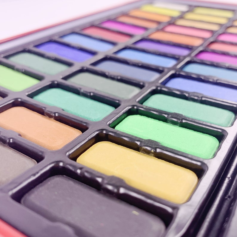 May include: A close-up of a watercolor paint set with a variety of colors in individual pans. The pans are arranged in rows and are set in a black plastic tray.