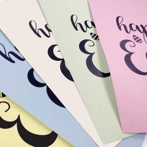 May include: A close-up of four pastel-colored cards with the words "happy Easter" printed in black script. The cards are light yellow, blue, green, and pink. The "E" in Easter is decorated with a bee.
