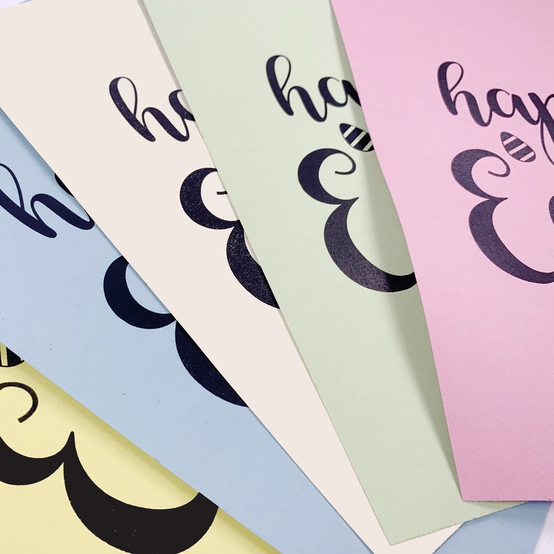 May include: A close-up of four pastel-colored cards with the words "happy Easter" printed in black script. The cards are light yellow, blue, green, and pink. The "E" in Easter is decorated with a bee.