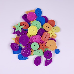 May include: A collection of colourful plastic buttons in various shapes, including stars, circles, squares, and rectangles. The buttons are arranged in a pile on a white background.