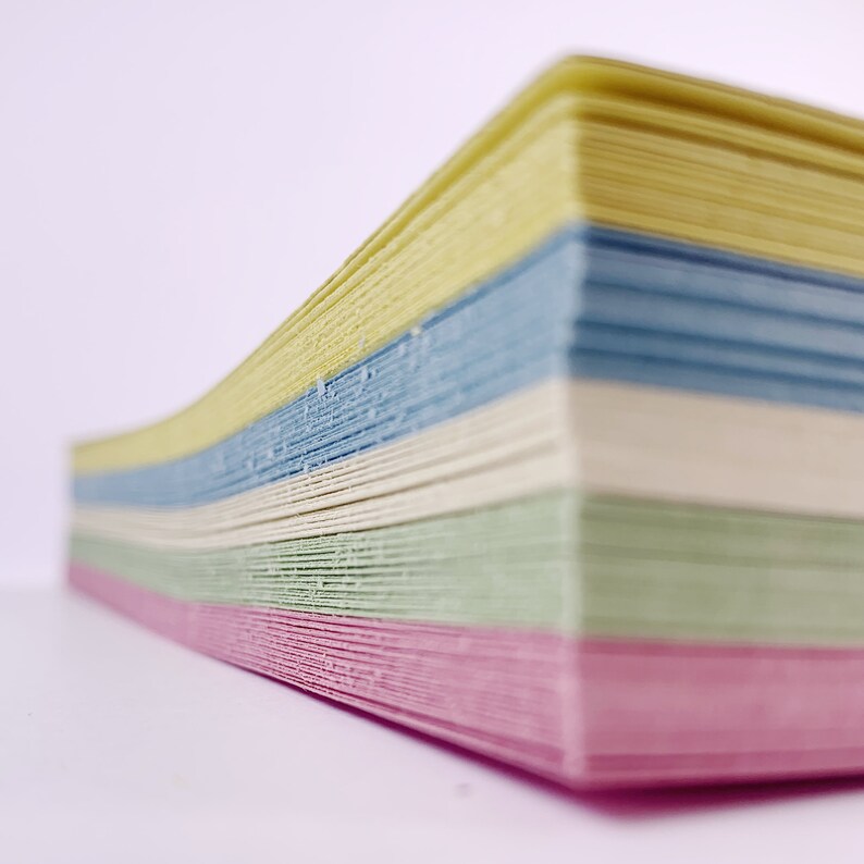 May include: A stack of colorful paper sheets in pastel shades of pink, green, cream, blue, and yellow. The paper is stacked in a slightly curved shape, with the edges of the sheets visible. The background is plain white.