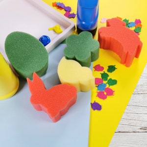 May include: A set of colourful foam stamps in various shapes, including a flower, a bunny, a heart, and a butterfly. The stamps are on a yellow surface with a white tray and a blue paint bottle.
