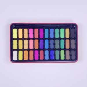 May include: A rectangular tin box filled with 36 watercolor paint pans. The pans are arranged in rows and are a variety of colors, including red, orange, yellow, green, blue, purple, and brown. The tin box has a black lid with a red border.