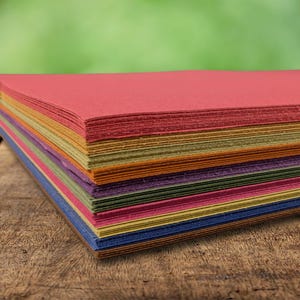 May include: A stack of rectangular sheets of leather in various colours including red, yellow, orange, purple, green, and blue. The sheets are layered on top of each other, showing the textured edges and the thickness of each individual piece of material.