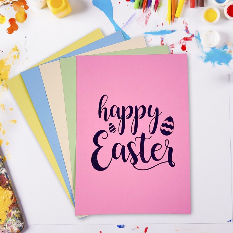 May include: A stack of pastel-colored cards with a pink card on top. The pink card has the words "happy Easter" in a dark blue script with two Easter egg illustrations. Paint and art supplies are in the background.