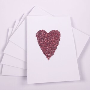 Puede incluir: Una pila de tarjetas blancas en blanco con un dise&ntilde;o de coraz&oacute;n rojo de purpurina en la tarjeta superior.