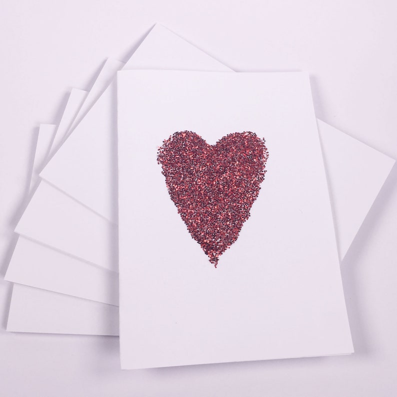 Puede incluir: Una pila de tarjetas blancas en blanco con un dise&ntilde;o de coraz&oacute;n rojo de purpurina en la tarjeta superior.
