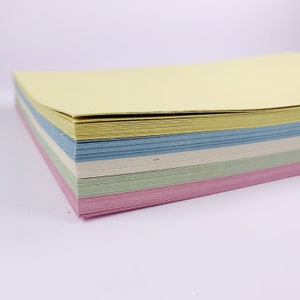 May include: A stack of pastel-colored paper sheets. The stack includes sheets in yellow, blue, white, green, and pink. The paper is stacked neatly, with the edges aligned. The paper appears to be new and unused.