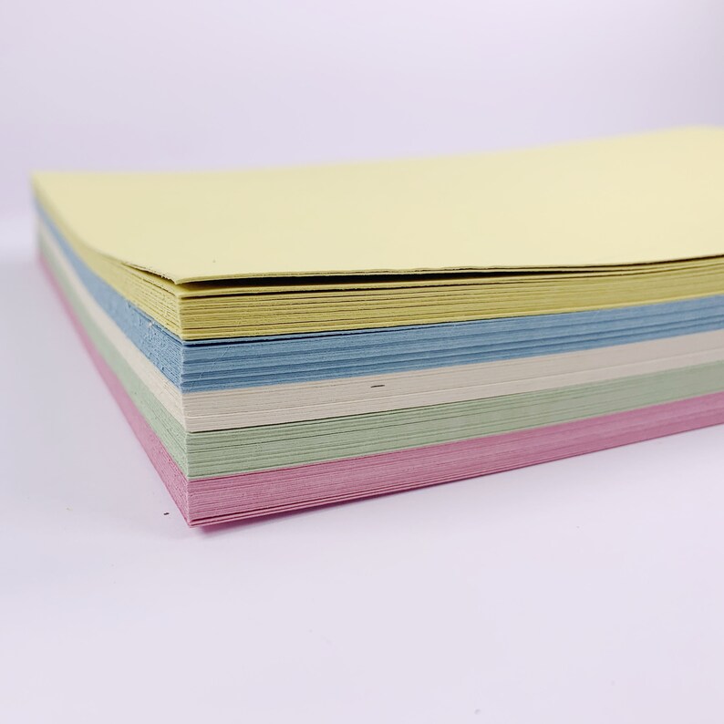 May include: A stack of pastel-colored paper sheets. The stack includes sheets in yellow, blue, white, green, and pink. The paper is stacked neatly, with the edges aligned. The paper appears to be new and unused.