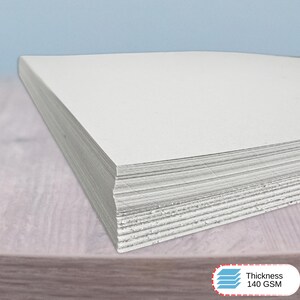 May include: A stack of rectangular white paper sheets with a smooth, matte finish. The paper is stacked neatly, showing the layered edges of the individual sheets. A small white label in the corner displays the text Thickness 140 GSM.