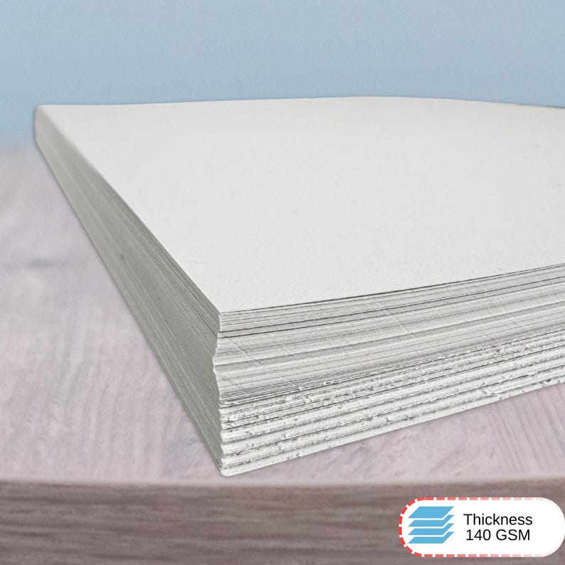 May include: A stack of rectangular white paper sheets with a smooth, matte finish. The paper is stacked neatly, showing the layered edges of the individual sheets. A small white label in the corner displays the text Thickness 140 GSM.