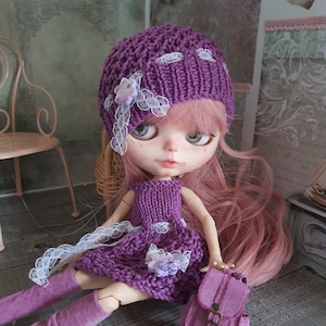Può includere: Una bambola di lana viola con i capelli rosa che indossa un cappello e un vestito di lana viola. La bambola tiene una piccola borsa viola.