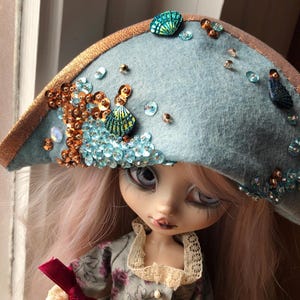 Könnte beinhalten: Hellblauer Filzhut mit goldenem Rand, verziert mit Pailletten und muschelförmigen Verzierungen. Der Hut ist Teil eines Puppenkostüms, mit rosa Haaren und einem geblümten Kleid.