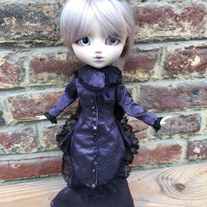 May include: A doll with pale skin, large grey eyes, and light grey hair, wearing a dark purple long-sleeved dress with a high collar, black lace trim, and a row of small buttons down the front, topped with a small black lace headpiece.