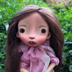 May include: A doll with long brown hair, large dark eyes, and rosy cheeks. The doll features a small mouth with a visible tooth and wears a pink dress with a ruffled tulle collar and a white fabric accent. A small white floral hair accessory is visible.