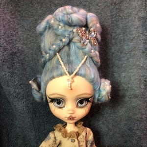 Può includere: Una bambola con capelli blu e bianchi acconciati in un'elaborata acconciatura con accenti di perle. La bambola ha grandi occhi blu, una carnagione chiara e indossa un vestito beige con un motivo floreale.