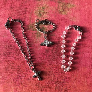 May include: Three pieces of jewellery on a red surface. One is a silver chain necklace with a cross pendant. Another is a silver ring with a heart-shaped chain and a winged heart charm. The third is a beaded bracelet.
