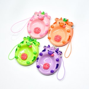 May include: Four cow-shaped charms with glossy finishes in pink, orange, lime green, and purple. Each charm features a central indentation, a small flower-like detail, and a matching colored loop strap attached to a metal ring for hanging purposes.
