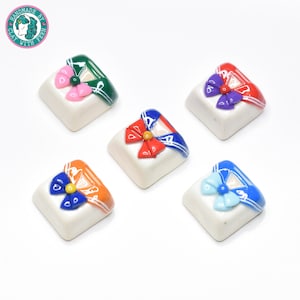 Puede incluir: Cinco teclas de diferentes colores con un diseño inspirado en Sailor Moon. Cada tecla tiene una base blanca con un cuello azul y un lazo de colores. Los lazos son rojo, rosa, morado, naranja y azul.