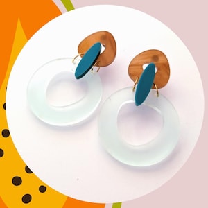 May include: A pair of contemporary earrings with a translucent, pale blue circular design. Each earring has a brown, oval-shaped accent and a teal blue, elongated oval accent. The earrings are set against a white background.