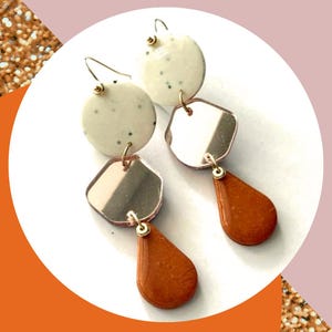 May include: A pair of dangle earrings with a layered design. The earrings feature a white speckled circle, a mirrored geometric shape, and a teardrop-shaped pendant in shades of brown. Gold-colored hooks and accents complete the design.