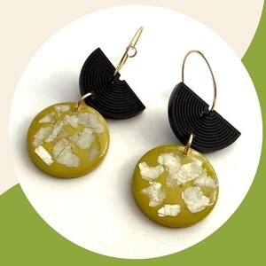 May include: A pair of dangle earrings featuring a black semi-circular component with a ribbed texture attached to a gold-toned wire hoop. Below, a circular mustard yellow pendant is embedded with irregular, reflective silver-toned foil flakes.