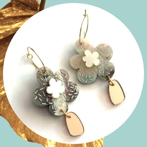 May include: A pair of hoop earrings featuring flower-shaped charms with a mottled green and grey surface, accented by small pink dash patterns. Each earring includes a small white flower centre and a dangling, smooth, light peach-coloured tab.