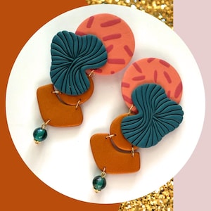 May include: A pair of handmade earrings with a modern design. The earrings feature orange, teal, and brown elements, including circular and leaf-shaped components. Each earring has a green bead at the bottom, suspended by a gold-colored chain.