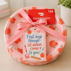 Könnte beinhalten: Ein Stapel runder Pappteller mit rosa Hintergrund und rotem Apfelmuster, zusammengebunden mit einem rosa Band. Eine rote DoorDash-Geschenkkarte und ein rechteckiges Etikett sind befestigt, mit dem Text First days through- let dinner come to you!