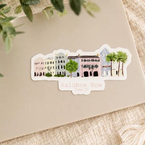 May include: A sticker featuring a watercolor illustration of Rainbow Row in Charleston, SC. The sticker depicts a row of colorful buildings with trees and the text "Rainbow Row" in a white banner.