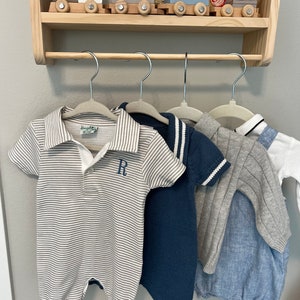 Puede incluir: Cuatro prendas de bebé colgadas en un estante de madera. La ropa incluye un mono a rayas con la letra "R" bordada en el pecho, un suéter azul con rayas blancas en las mangas, un suéter gris con rayas blancas en las mangas y un mono azul claro con cuello blanco.