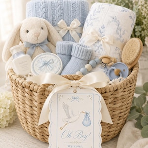 May include: A wicker basket containing baby essentials. It includes a blue cable-knit blanket, a soft toy bunny, and a tag with a stork illustration and the words "Oh Boy!". Also present are baby socks, a dummy, and a brush.