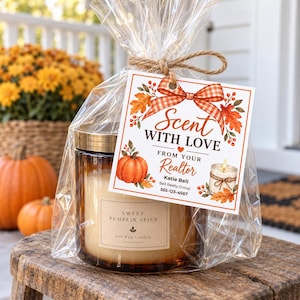 May include: A glass jar candle with a gold lid, labelled Sweet Pumpkin Spice, wrapped in clear plastic with a twine-tied tag. The tag features an orange plaid bow, autumn leaves, a pumpkin, and text: Scent With Love From Your Realtor, Katie Bell, Bell Realty Group, 555-123-4567.