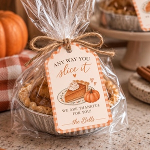 Puede incluir: Una pequeña tarta de calabaza en un molde redondo de aluminio, envuelta en plástico transparente y atada con cuerda. Una etiqueta rectangular con borde naranja a cuadros muestra el texto 'ANY WAY YOU slice it', 'WE ARE THANKFUL FOR YOU!' y 'the Bells'.